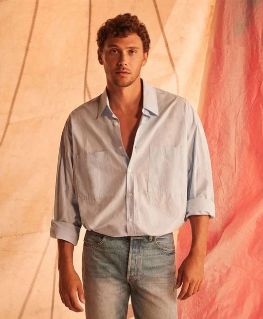Young adult man in light blue button-down and jeans poses against a textured peach background, portrait studio shot. Casual fashion.