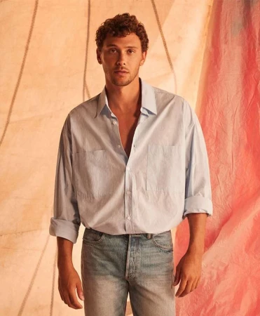 Young adult man in light blue button-down and jeans poses against a textured peach background, portrait studio shot. Casual fashion.