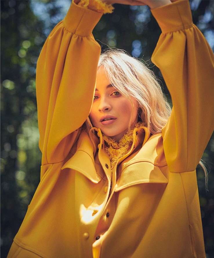 Portrait of a young woman with blond hair wearing a yellow coat, posing with her arms above her head against a blurred green background.