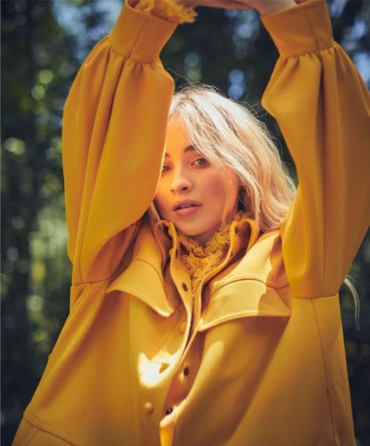 Portrait of a young woman with blond hair wearing a yellow coat, posing with her arms above her head against a blurred green background.