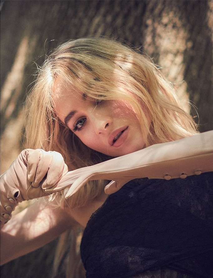 Close-up portrait of a blond woman in tan gloves and a black lace top, with a neutral facial expression, posing outdoors in soft lighting.