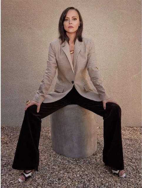 Portrait of Christina Ricci sitting on a concrete stool wearing a tan patterned blazer and dark brown flared pants in a photo studio.