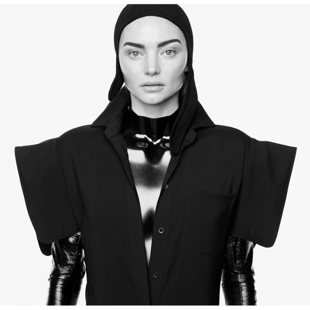 Monochrome portrait of an adult woman with a knit cap, wearing a button-down shirt and gloves, against a white backdrop.