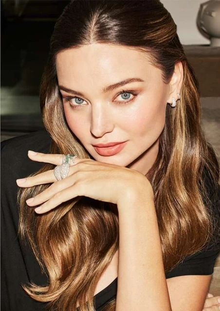 Close-up studio portrait of Miranda Kerr with fair skin, light eyes, and long brunette hair wearing a black top, posing and wearing rings.