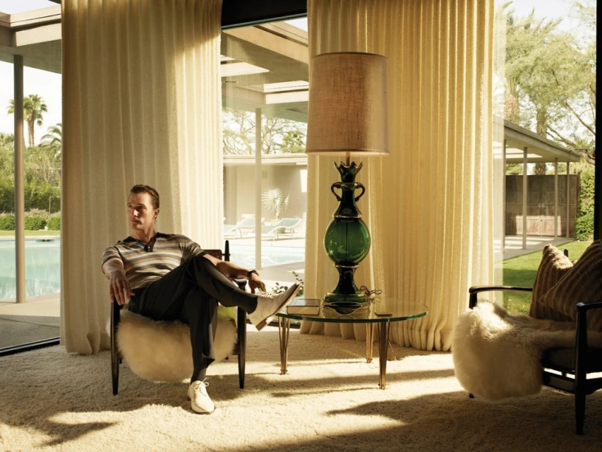 Man seated in chair inside mid-century modern home with floor-to-ceiling windows overlooking swimming pool and backyard.