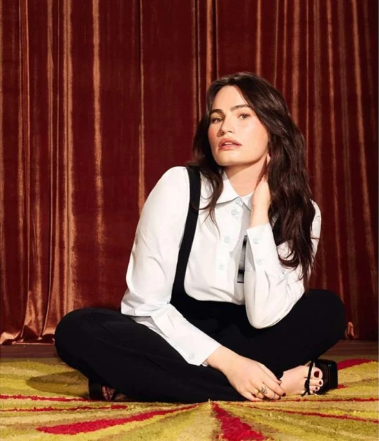 Portrait of a brunette woman with fair skin sitting cross-legged on an ornate rug in front of a red-brown velvet curtain backdrop.