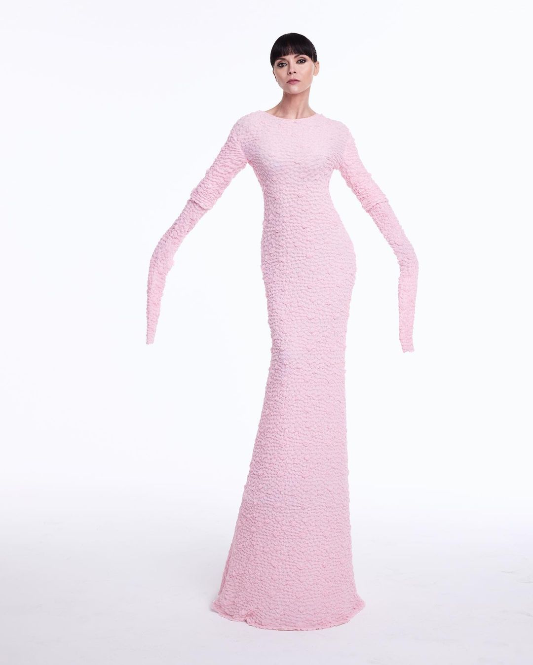 Elegant woman poses in a pink, textured gown against a white background, creating a clean, high-fashion studio shot with minimalist appeal.
