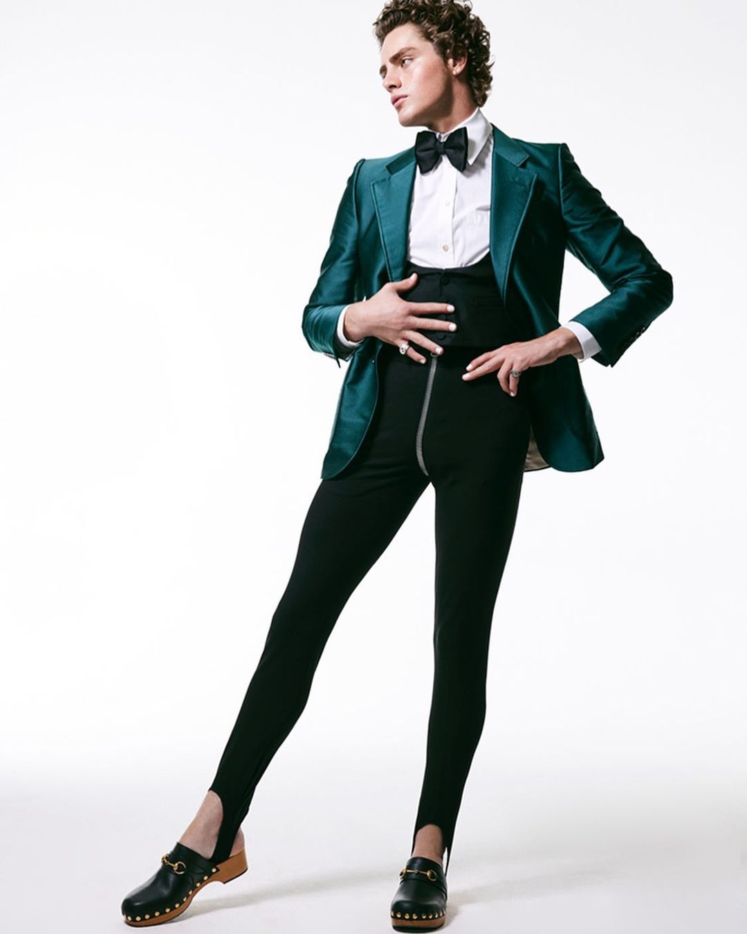 A male fashion model poses in a green velvet suit jacket, white shirt, bow tie, high-waisted leggings, and wooden clogs in a studio shot.