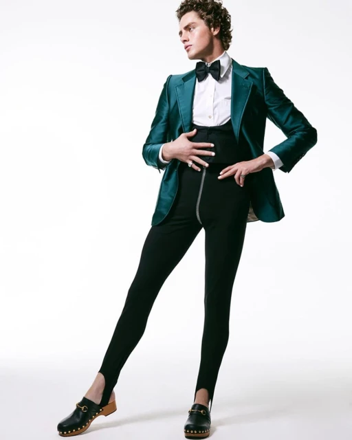 A male fashion model poses in a green velvet suit jacket, white shirt, bow tie, high-waisted leggings, and wooden clogs in a studio shot.