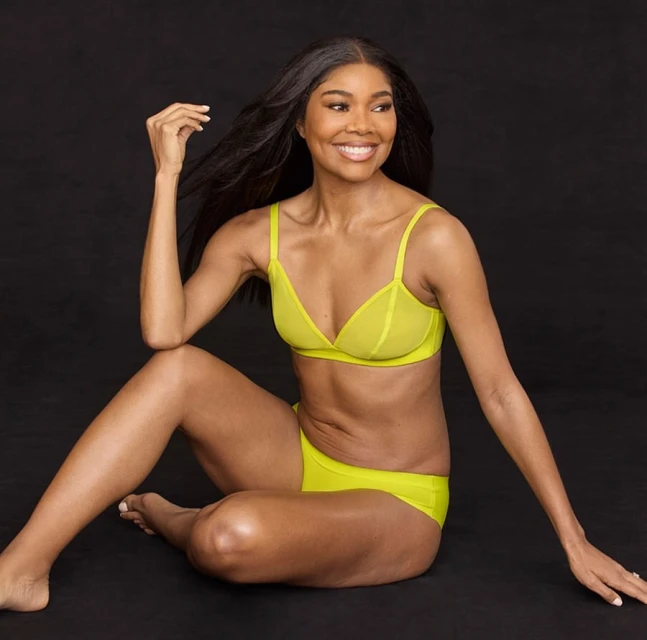 Adult woman poses in a seated position wearing a chartreuse lingerie set in front of a black studio backdrop with soft lighting.