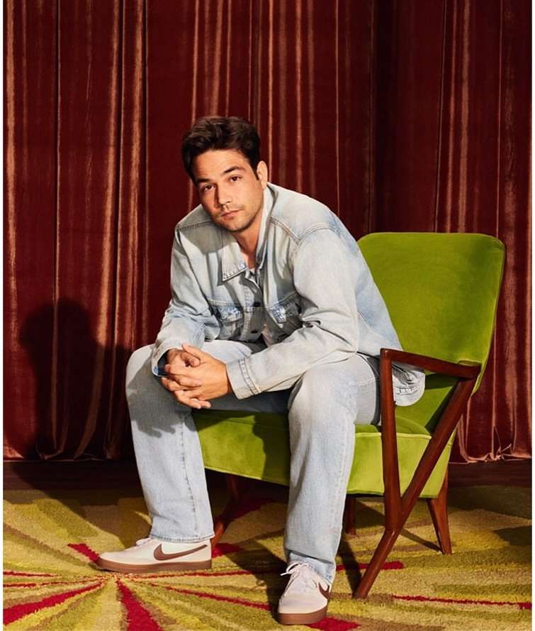 Man in light denim jacket and jeans seated in a green armchair against red velvet curtains by Nino Munoz