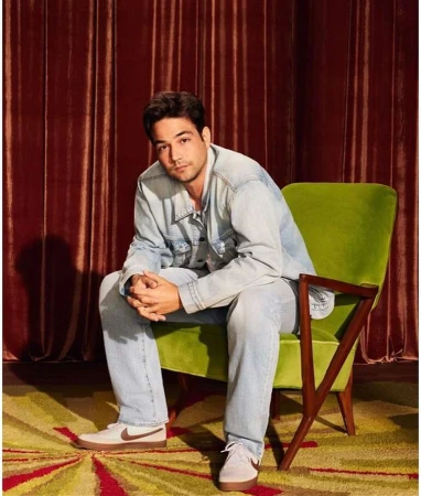 Man in light denim jacket and jeans seated in a green armchair against red velvet curtains by Nino Munoz