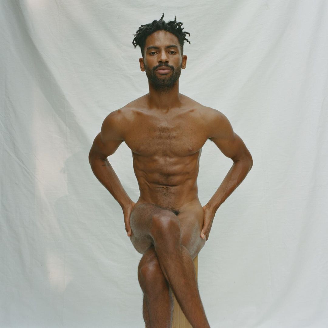 Nude African-American male seated with arms on thighs against white backdrop, showcasing fitness and physique in studio lighting.