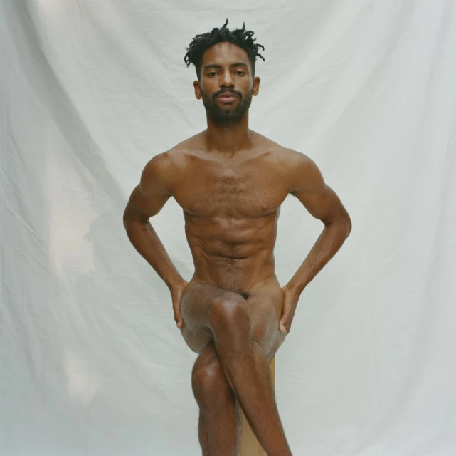 Nude African-American male seated with arms on thighs against white backdrop, showcasing fitness and physique in studio lighting.