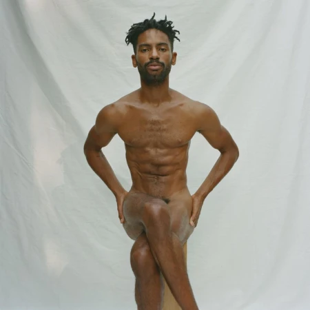 Nude African-American male seated with arms on thighs against white backdrop, showcasing fitness and physique in studio lighting.