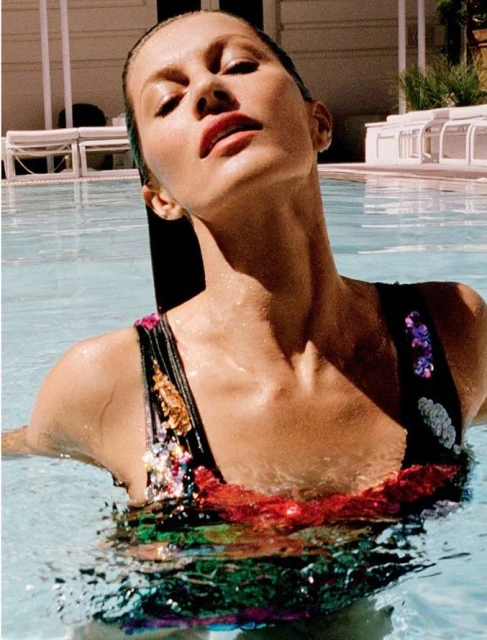 A woman in a colorful swimsuit with sunlit skin is partially submerged in a blue swimming pool, looking up with a relaxed facial expression.