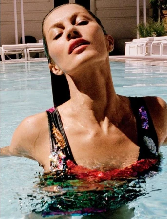 A woman in a colorful swimsuit with sunlit skin is partially submerged in a blue swimming pool, looking up with a relaxed facial expression.
