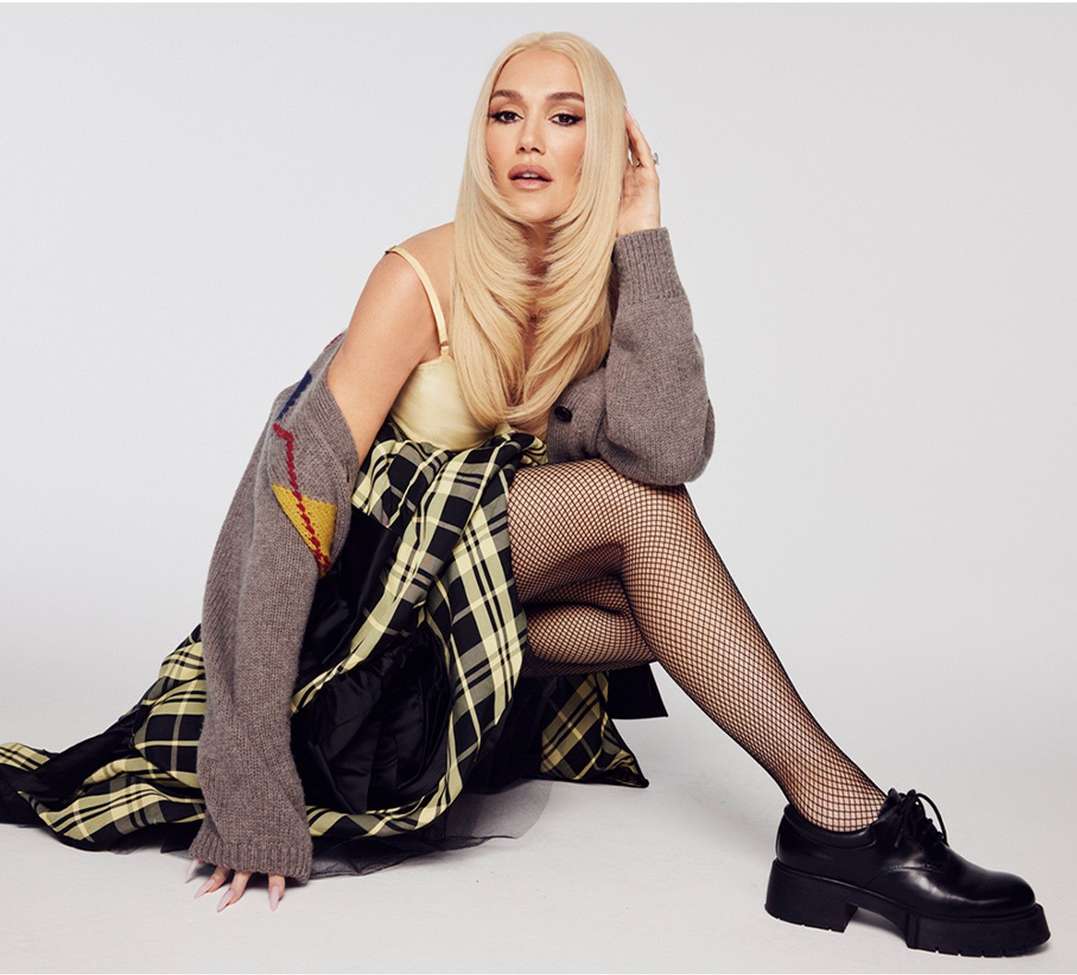 Gwen Stefani with long blonde hair seated against a plain white background wearing fishnet tights and a plaid skirt.