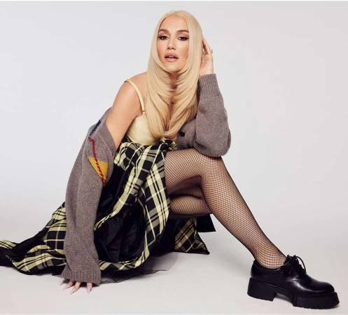 Gwen Stefani with long blonde hair seated against a plain white background wearing fishnet tights and a plaid skirt.
