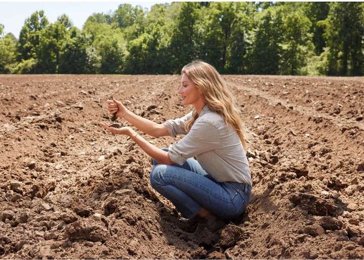 Woman squats in a farm field, holding soil in her hands, wearing jeans and a light brown shirt, with trees in the background on a sunny day.