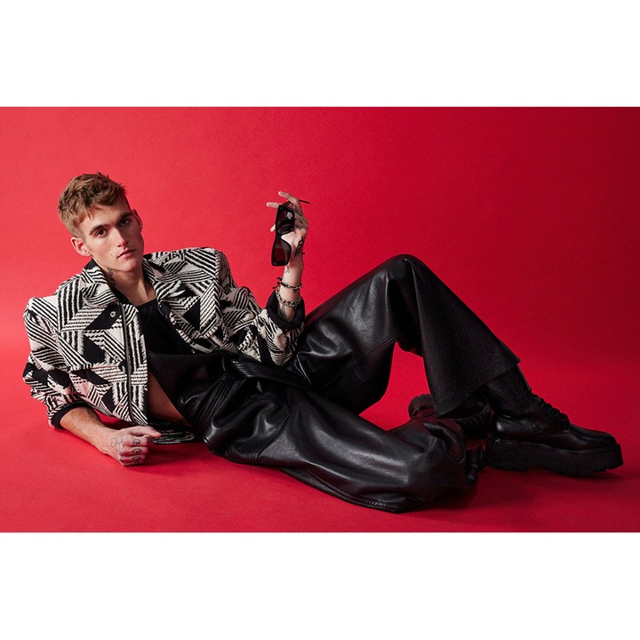 Adult male poses on red background wearing a black and white patterned jacket, black leather pants and boots, and holding sunglasses.