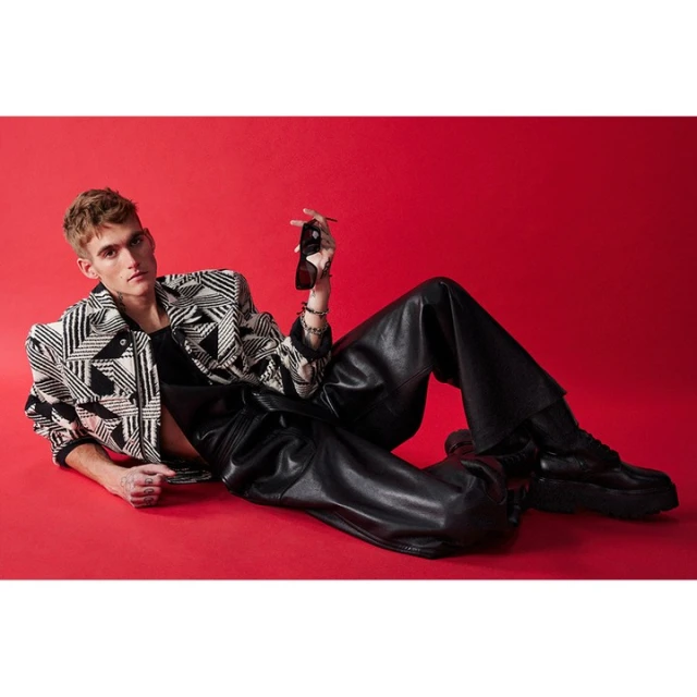 Adult male poses on red background wearing a black and white patterned jacket, black leather pants and boots, and holding sunglasses.
