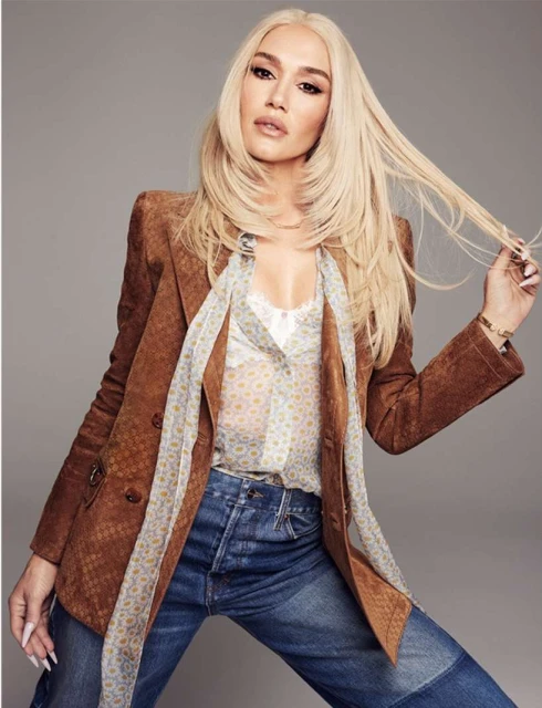 Blonde woman in a brown blazer and jeans poses in a studio. Her hair is long and straight and she is wearing a patterned blouse.