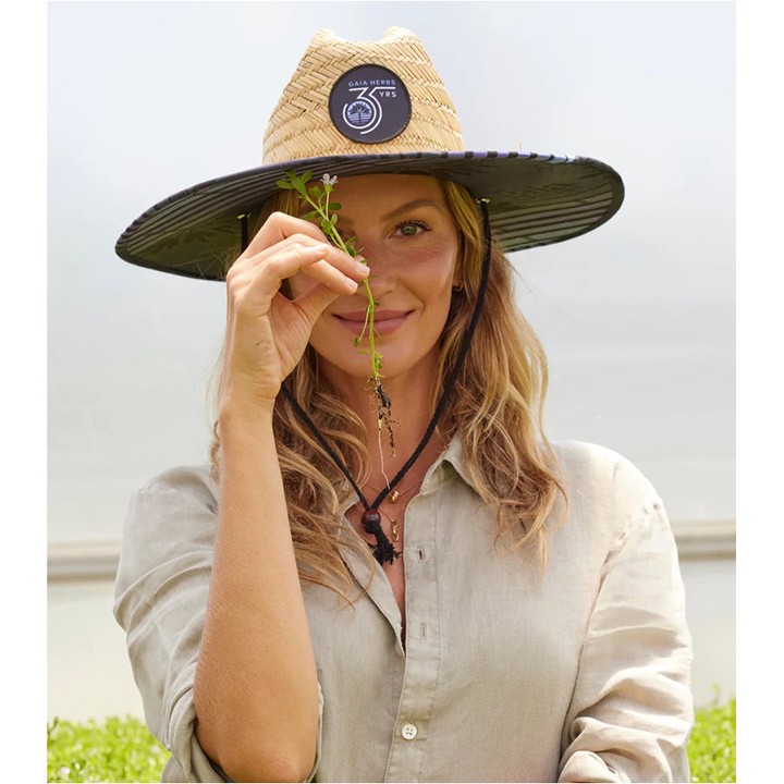 Adult woman in sunhat holding a small plant close to her face in an indoor greenhouse, wearing a button-down shirt. 