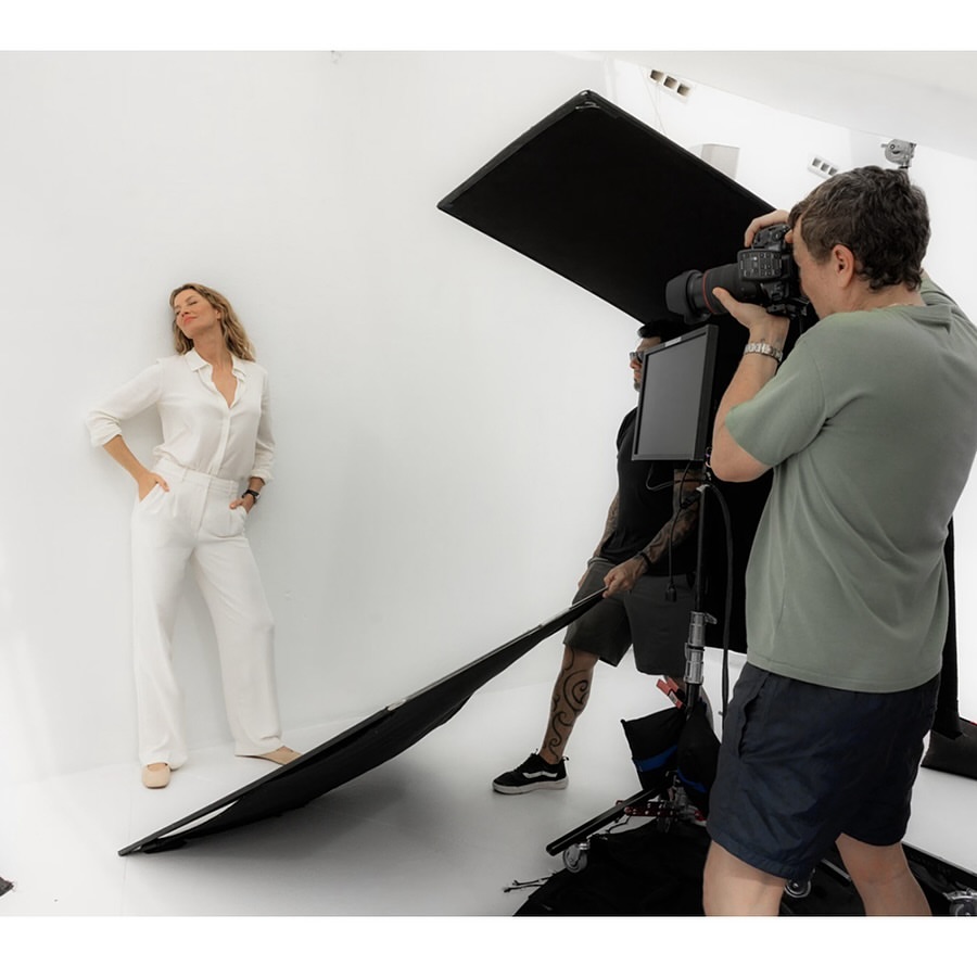 Fashion photo shoot in a studio with a woman posing in white pantsuit and a photographer and assistants with lighting equipment.