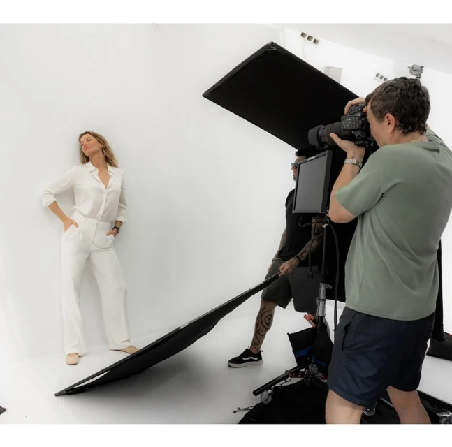 Fashion photo shoot in a studio with a woman posing in white pantsuit and a photographer and assistants with lighting equipment.