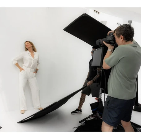 Fashion photo shoot in a studio with a woman posing in white pantsuit and a photographer and assistants with lighting equipment.