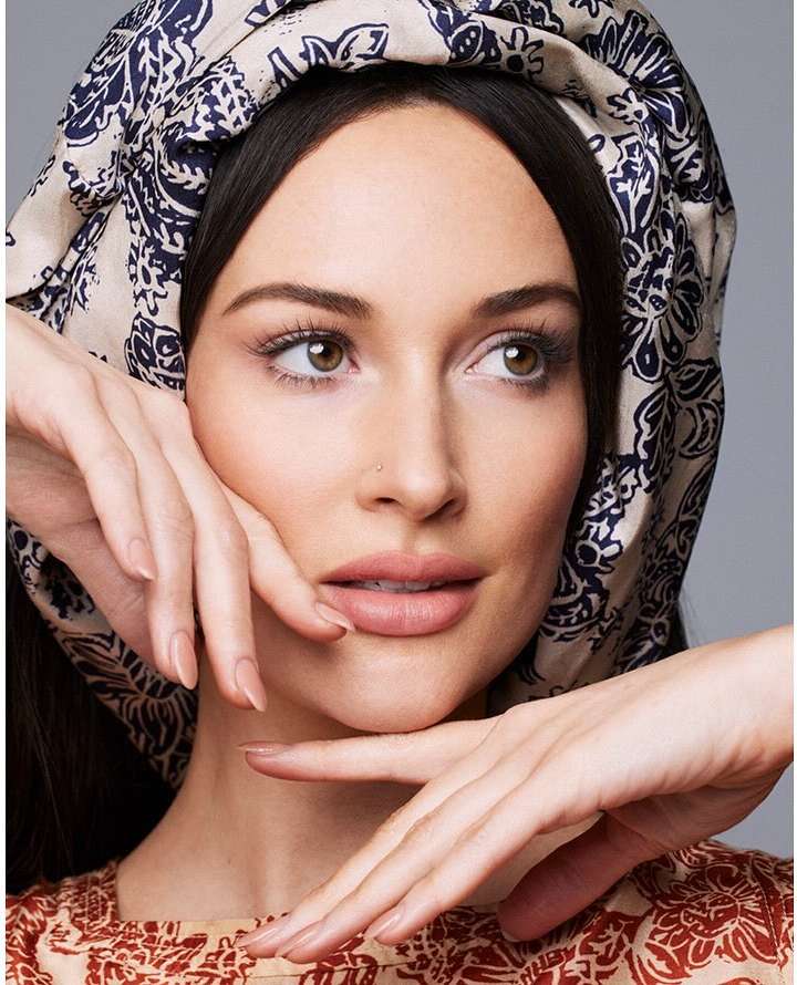 Close-up portrait of a woman with brown hair wearing a patterned headscarf and top, posing with her hands near her face.
