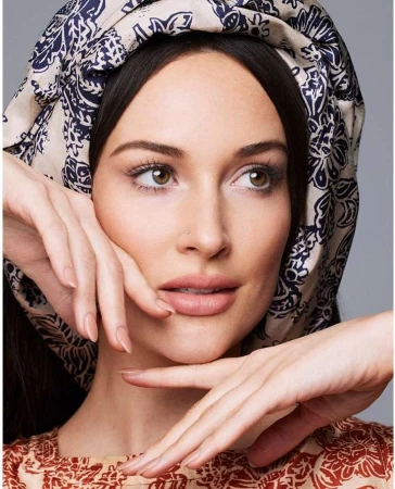 Close-up portrait of a woman with brown hair wearing a patterned headscarf and top, posing with her hands near her face.