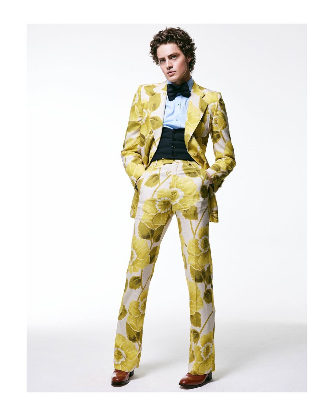 Man in a floral suit stands in a full shot on a white background. He wears a light blue dress shirt and a black bow tie.
