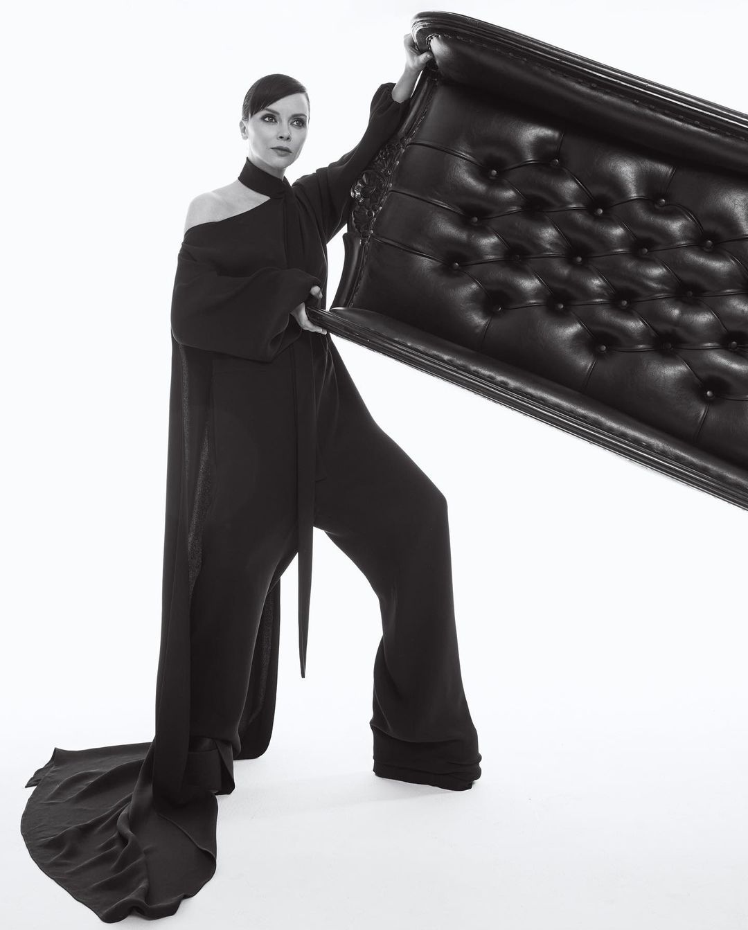 Adult woman in black, one-shoulder jumpsuit holding a tufted, black leather headboard in front of white backdrop in a studio environment.