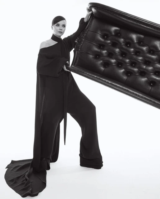 Adult woman in black, one-shoulder jumpsuit holding a tufted, black leather headboard in front of white backdrop in a studio environment.