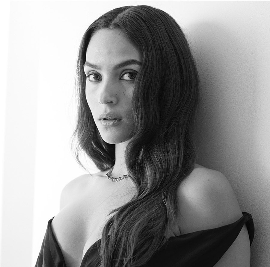 Black and white portrait of a woman with long dark wavy hair wearing an off-the-shoulder dark dress against a plain wall.