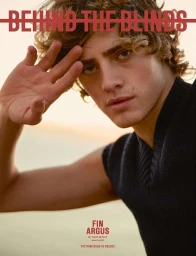 Portrait of young man with curly hair and blue eyes wearing a black sleeveless top, holding his hand up to his forehead against an outdoor sky