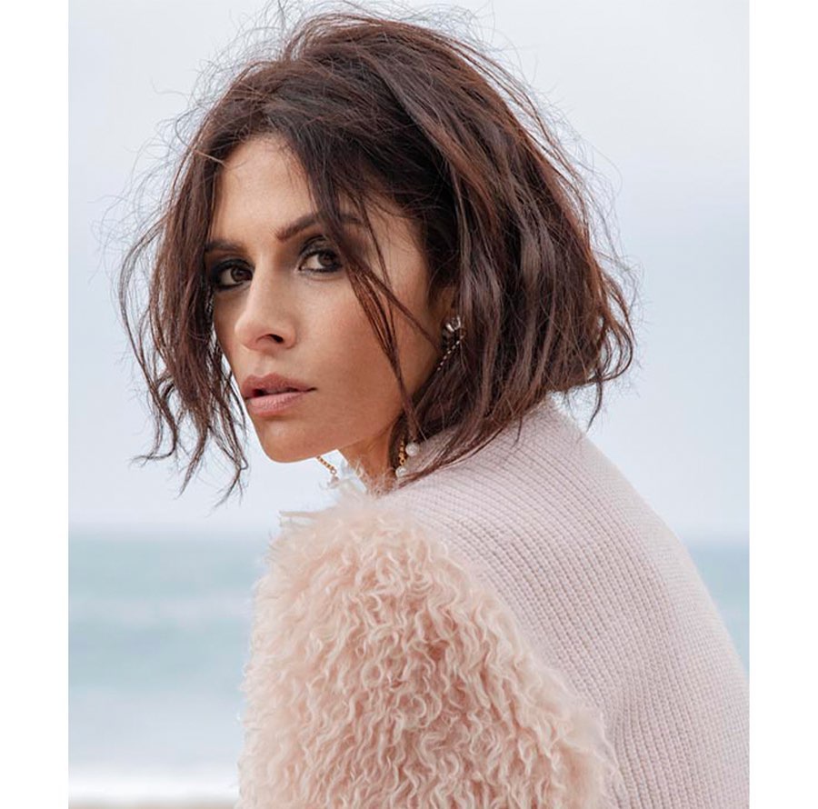 Portrait of a brunette woman in a light pink coat with a fuzzy texture, posing outdoors near a blurred beach and overcast sky.