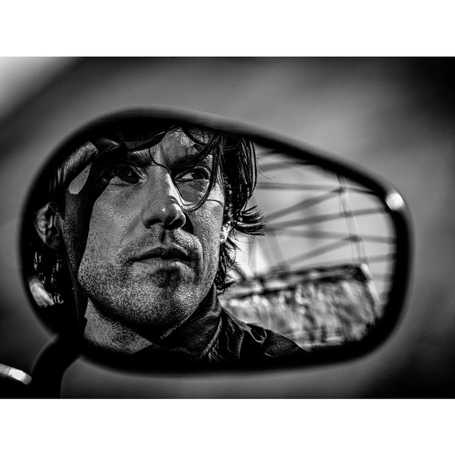 Black and white portrait of an adult male looking out from a motorcycle rearview mirror, capturing his face and the environment behind him.