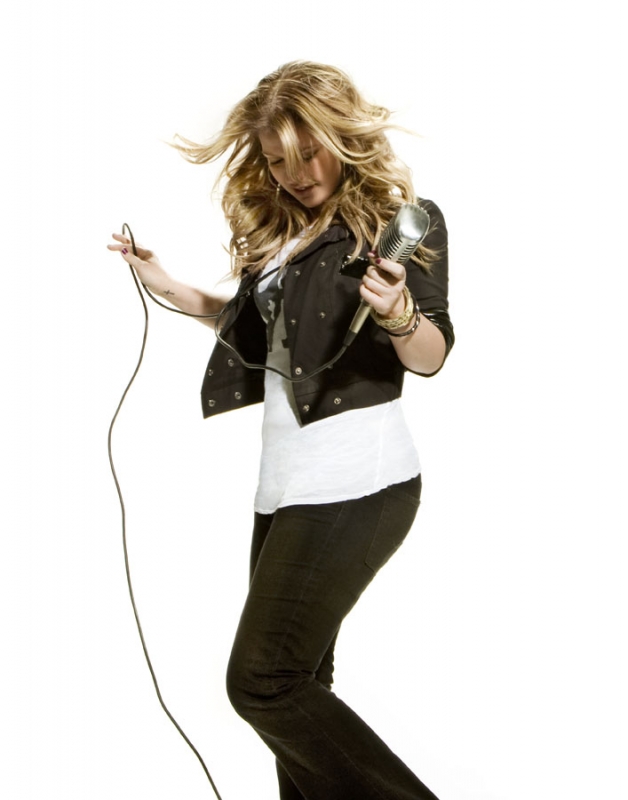 Blonde woman holding a vintage microphone and wire against a plain white background, wearing a dark jacket over a white top and dark pants