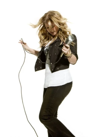 Blonde woman holding a vintage microphone and wire against a plain white background, wearing a dark jacket over a white top and dark pants