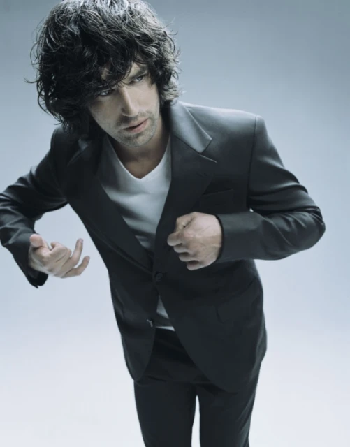 Portrait of a dark-haired man in a dark suit and white t-shirt, gazing sideways in a photo studio setting, medium shot, neutral expression.
