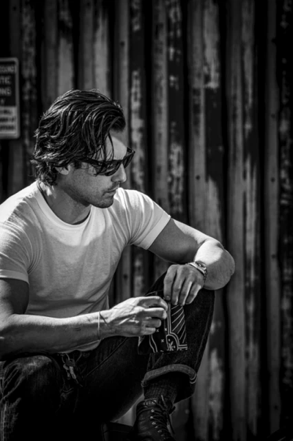 Monochrome portrait of a man in sunglasses and jeans squatting in front of a weathered metal wall, casually looking left.