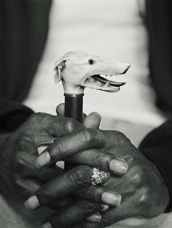 Close-up shot of an adult holding a cane with a carved dog head handle, set against a blurred background, captured in black and white.