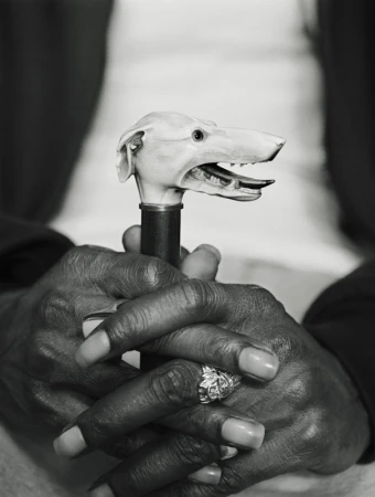 Close-up shot of an adult holding a cane with a carved dog head handle, set against a blurred background, captured in black and white.