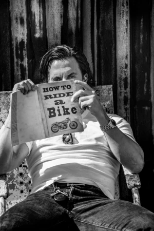 Man holding a book titled How to Ride a Bike while sitting in front of a corrugated metal wall, captured in high-contrast black and white.