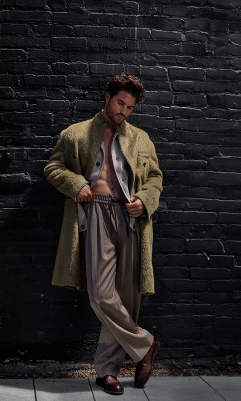 An adult male model poses against a dark brick wall, wearing a coat and beige pants with brown leather shoes in a full shot.