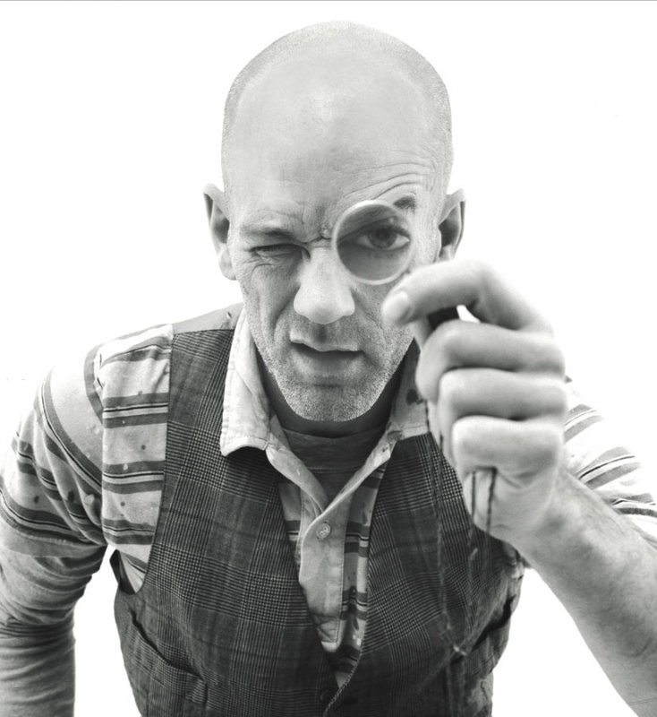 Black and white portrait of a mature adult male winking while looking through a monocle, wearing a vest and collared shirt.