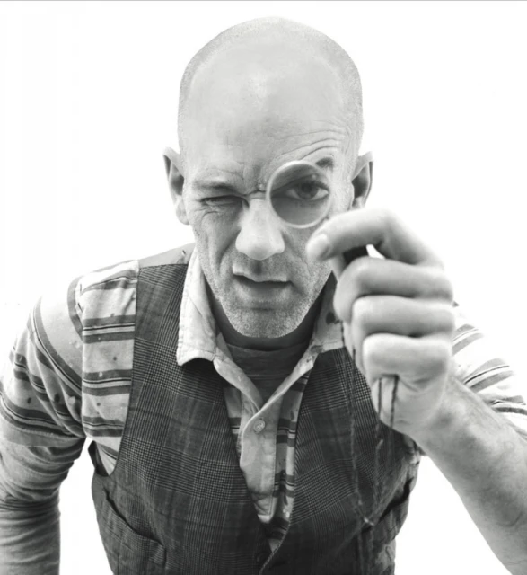 Black and white portrait of a mature adult male winking while looking through a monocle, wearing a vest and collared shirt.